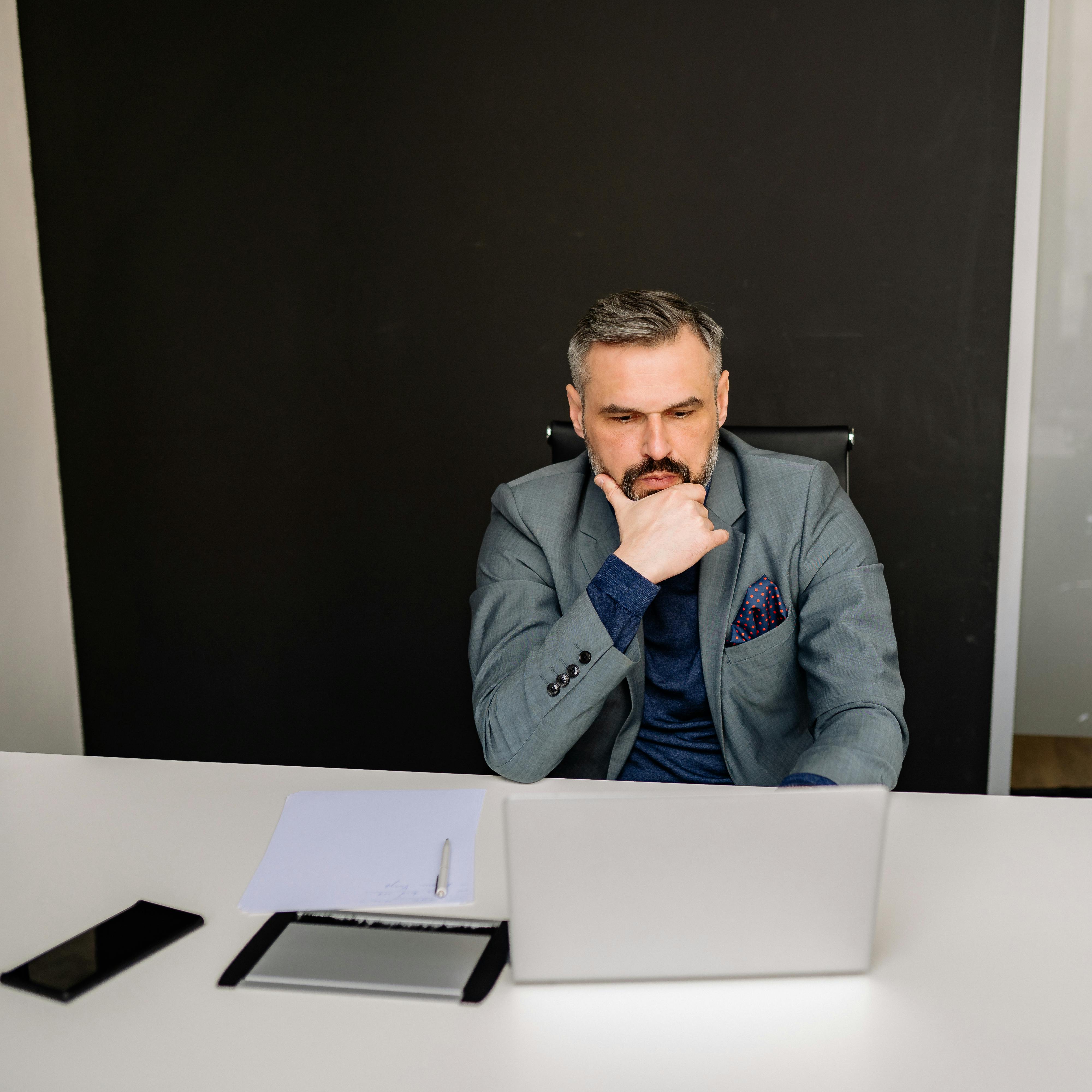 A thoughtful businessman in a suit working on his laptop in a modern office setting.