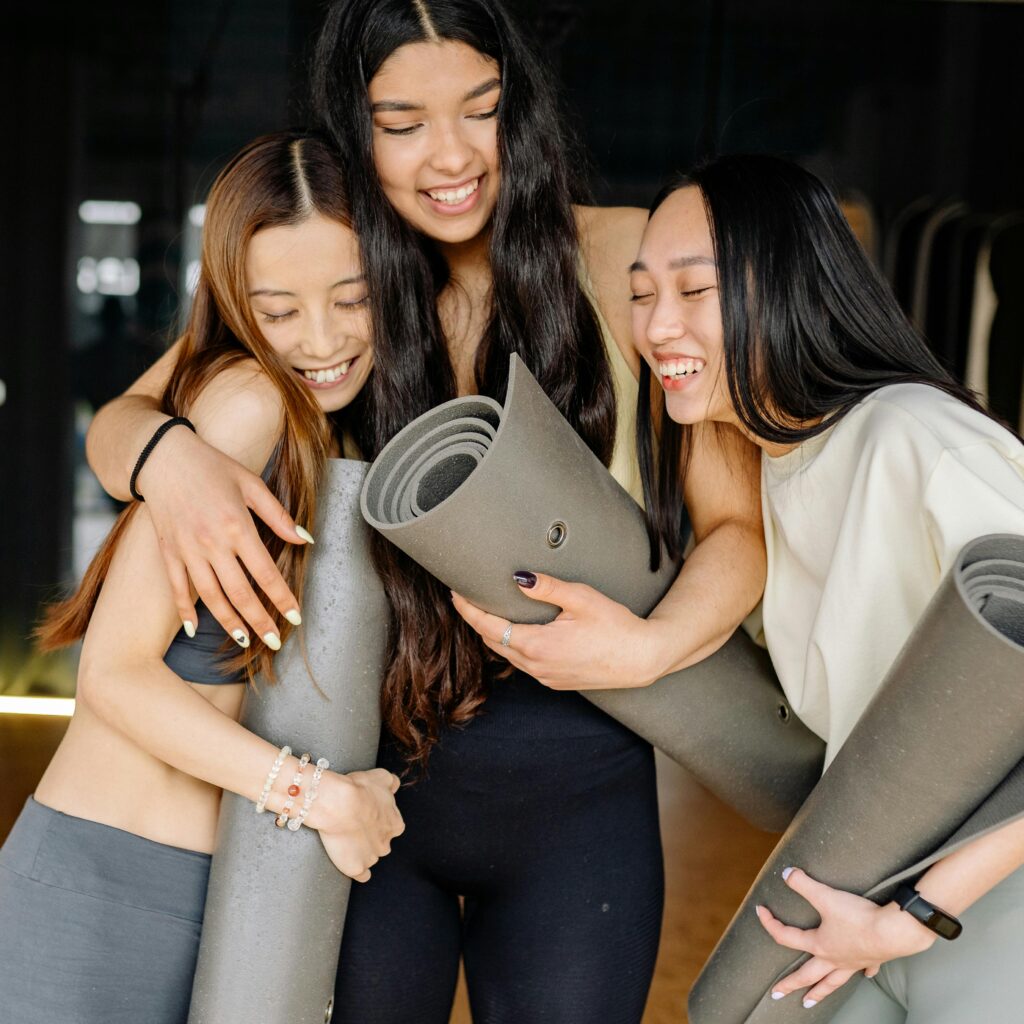 Three young women joyfully hugging and holding yoga mats, symbolizing friendship and healthy living.