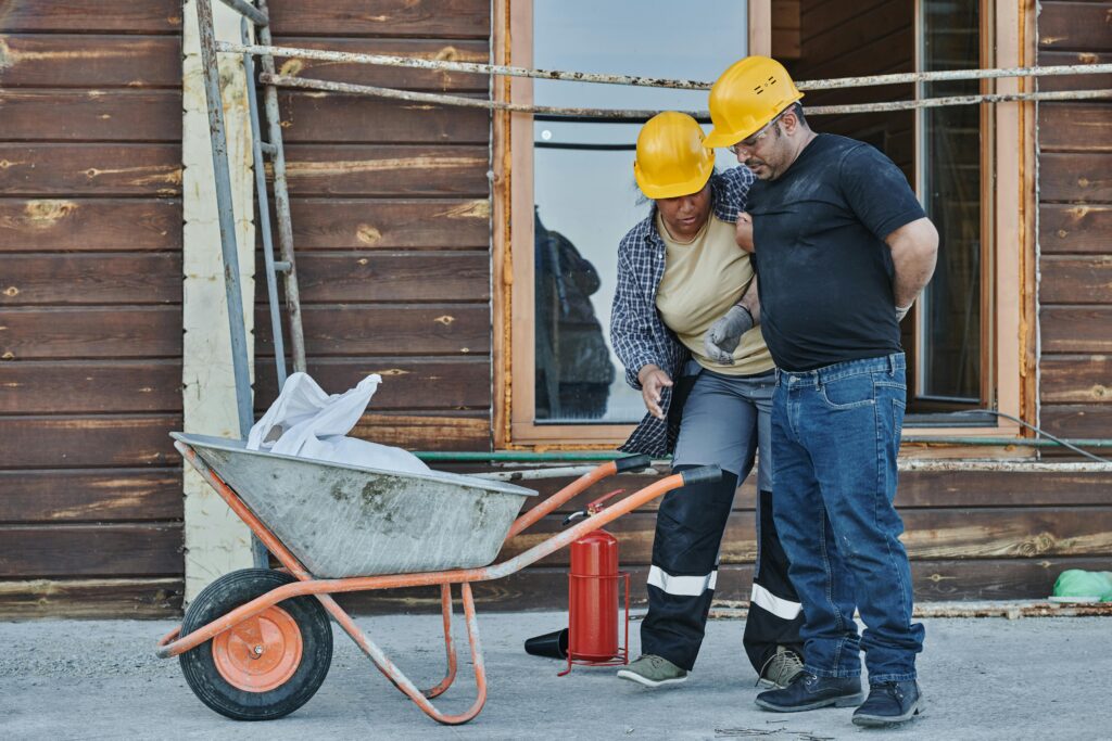 Two construction workers with hard hats assist an injured team member, highlighting workplace safety.